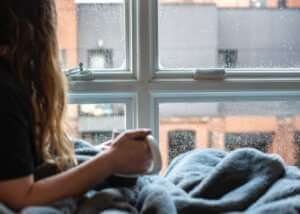 woman in gray sweater sitting beside window in relaxing bedroom
